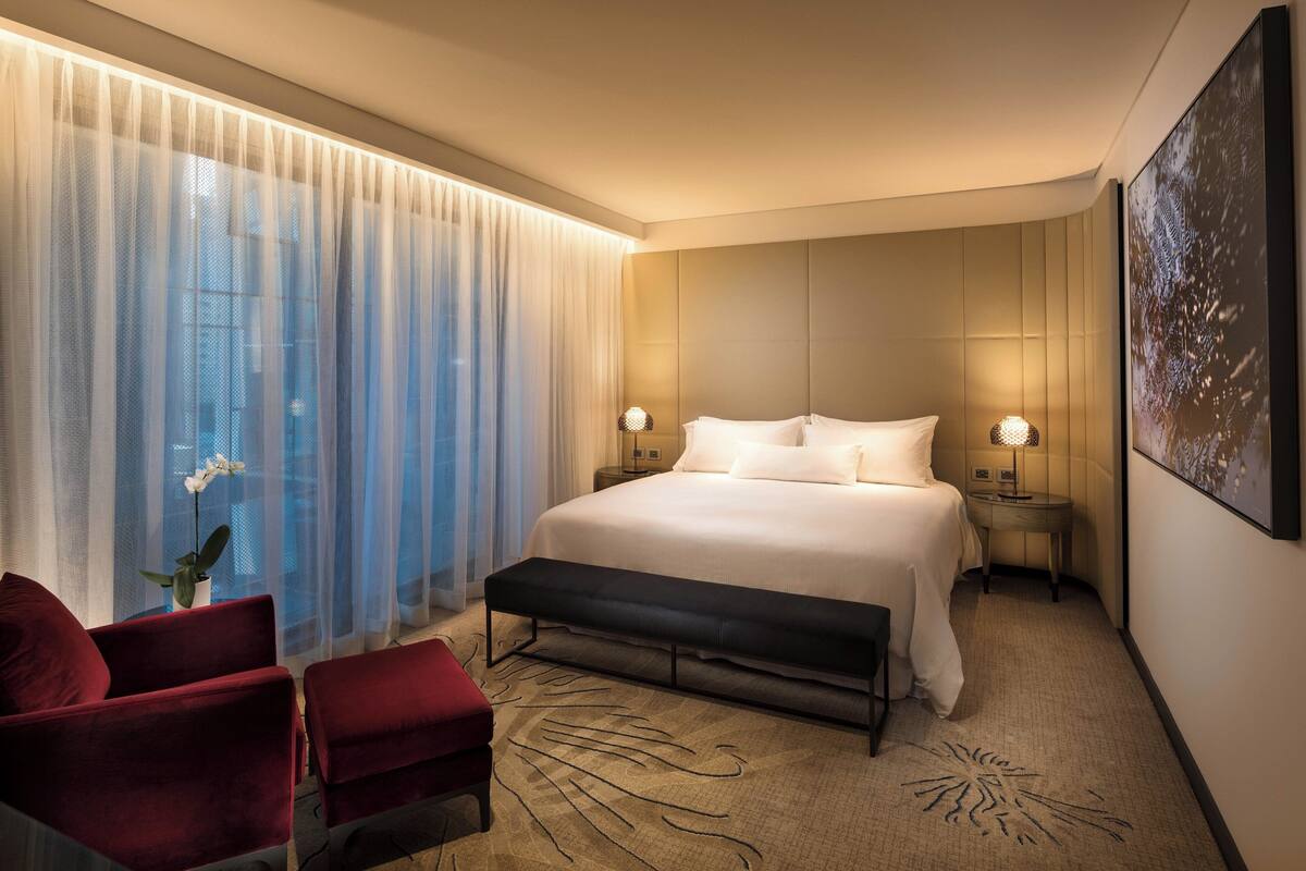 A modern hotel room with a large bed, two bedside lamps, a bench, a red chair, and a large window with sheer curtains.