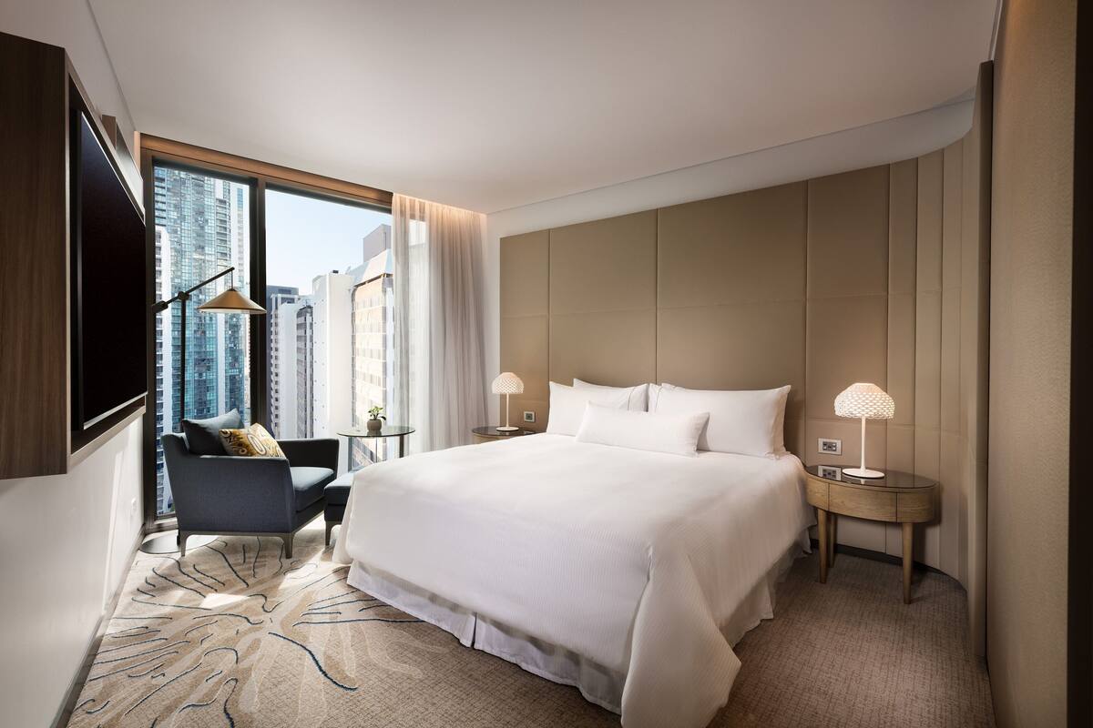 A modern hotel room with a large bed, a sitting area with a chair, a nightstand with a lamp, and a view of the city through large windows.