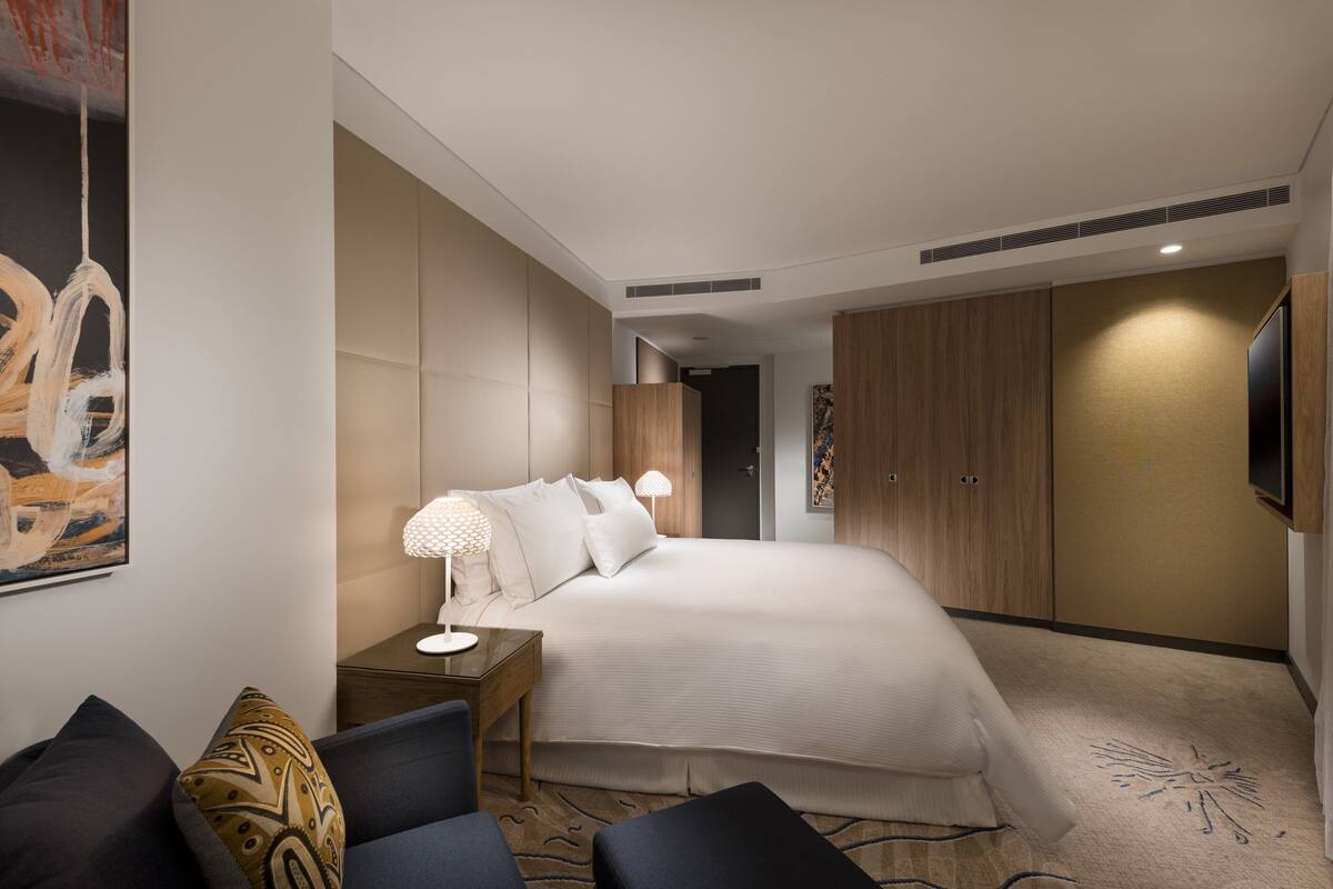 A modern hotel room with a large bed, a bedside table with a lamp, a chair, and a wardrobe.