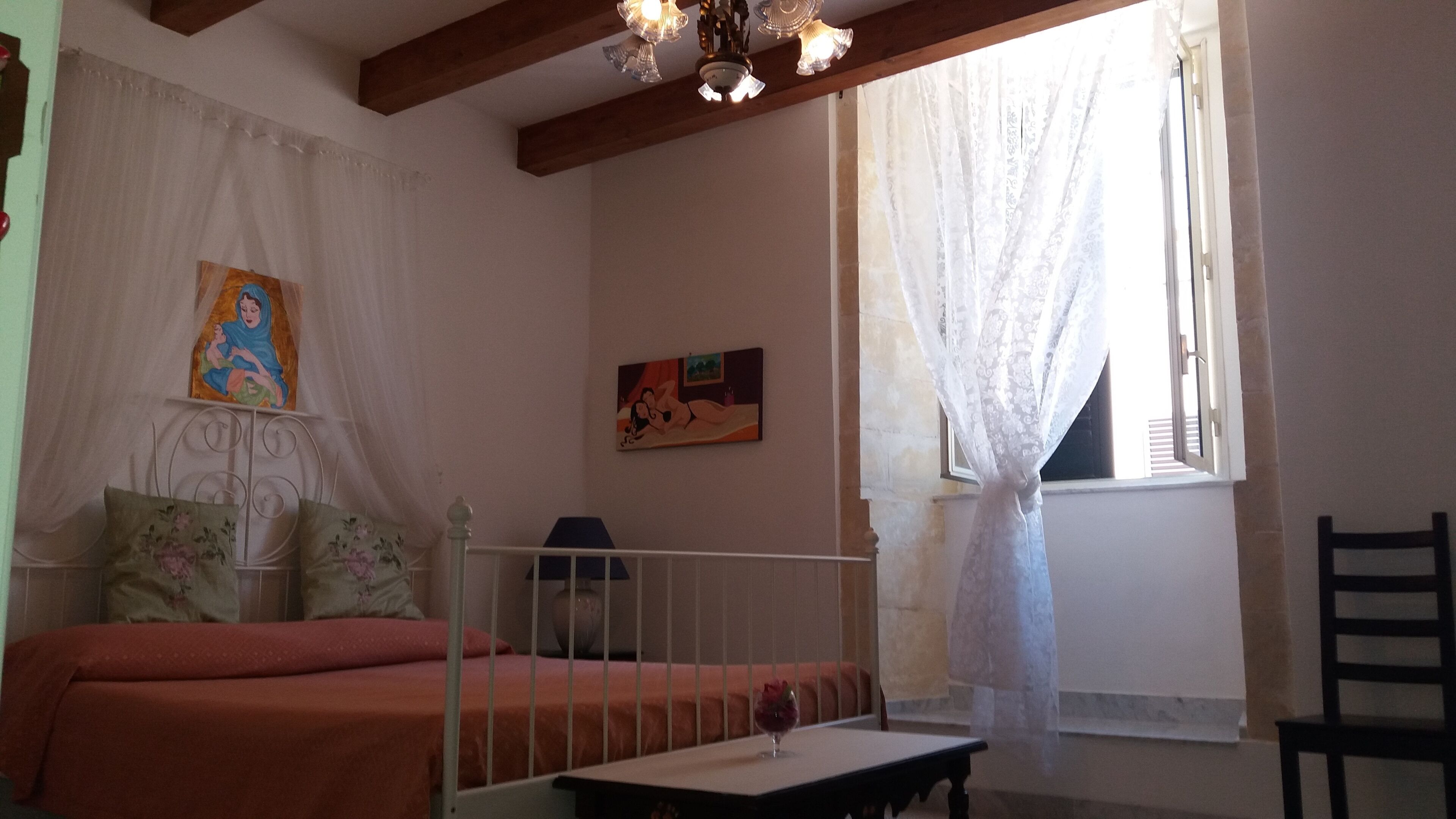 A bedroom with a bed, a small table, a chair, a window with curtains, and two wall-mounted paintings.