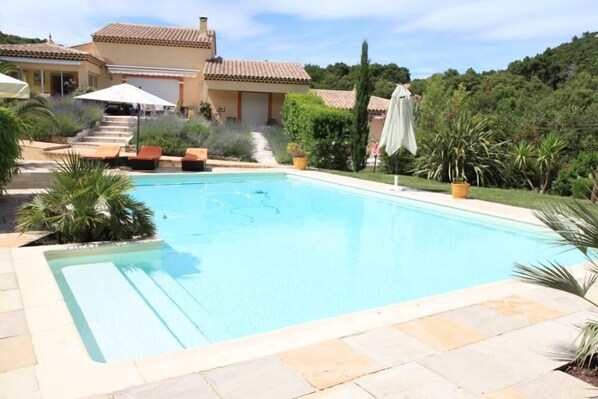 A swimming pool with lounge chairs and umbrellas, surrounded by greenery and a building with a tiled roof.