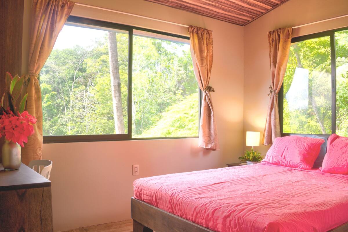 Une chambre à coucher avec un lit en bois, une literie rose et une vue sur la verdure grâce à de grandes fenêtres.