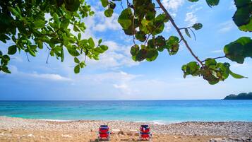A beach with two red beach chairs and a table under a tree, overlooking the sea.