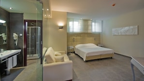 Una habitación de hotel moderna con ducha de vidrio, un sofá, una cama doble y una mesita de noche.