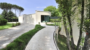 Un edificio moderno con fachada curva, camino de entrada pavimentado y un jardín bien cuidado con césped y árboles.
