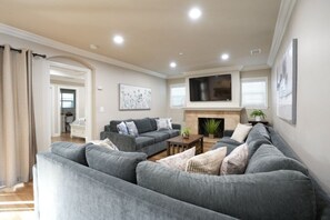 A living room with a grey sofa set, a flat-screen TV mounted above a fireplace, and a painting on the wall.