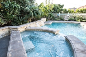 A swimming pool with a waterfall feature, surrounded by lush greenery and a white fence.