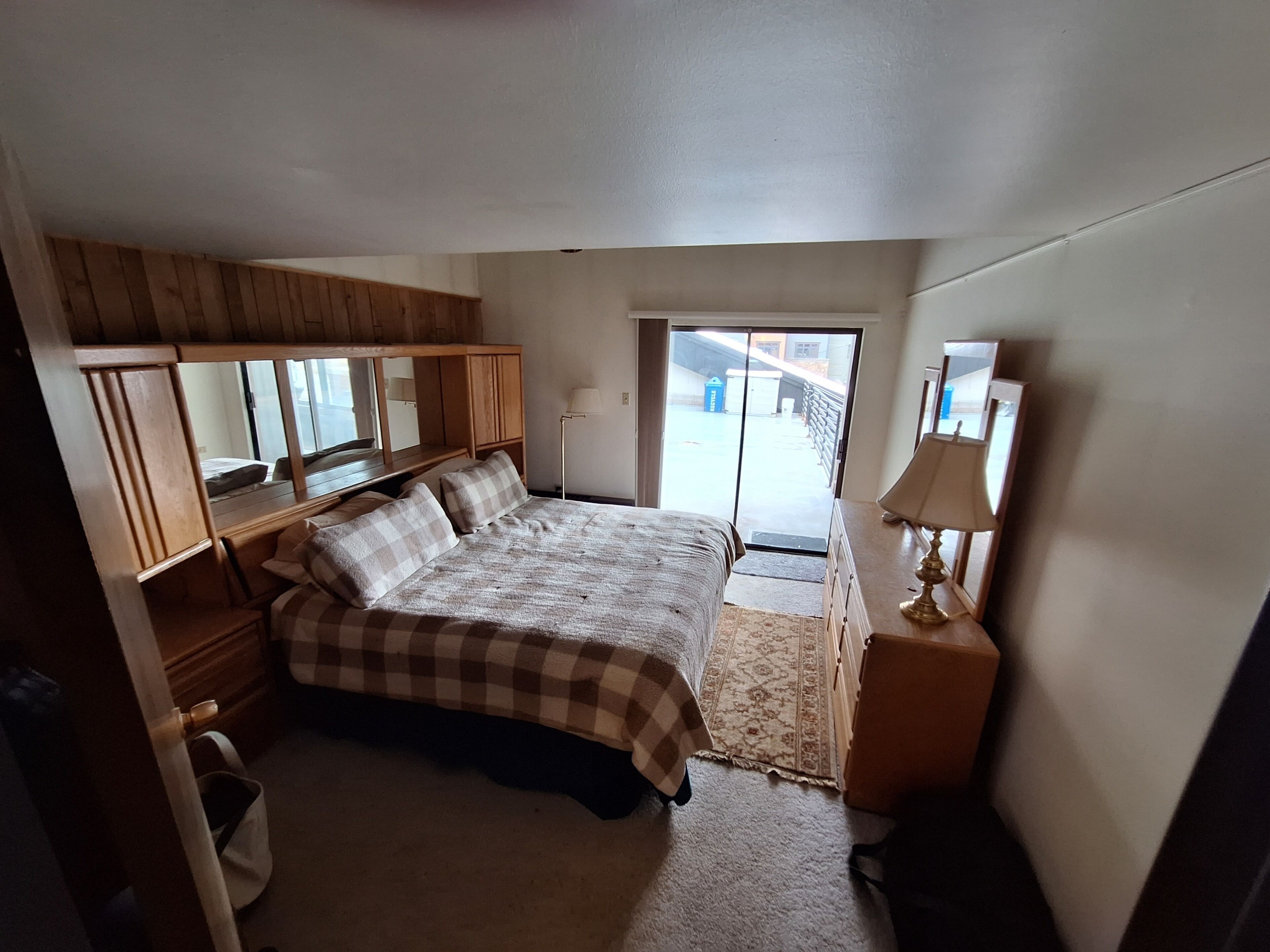 A bedroom with a bed, wooden wardrobe, a nightstand, a lamp, and a view through a door.
