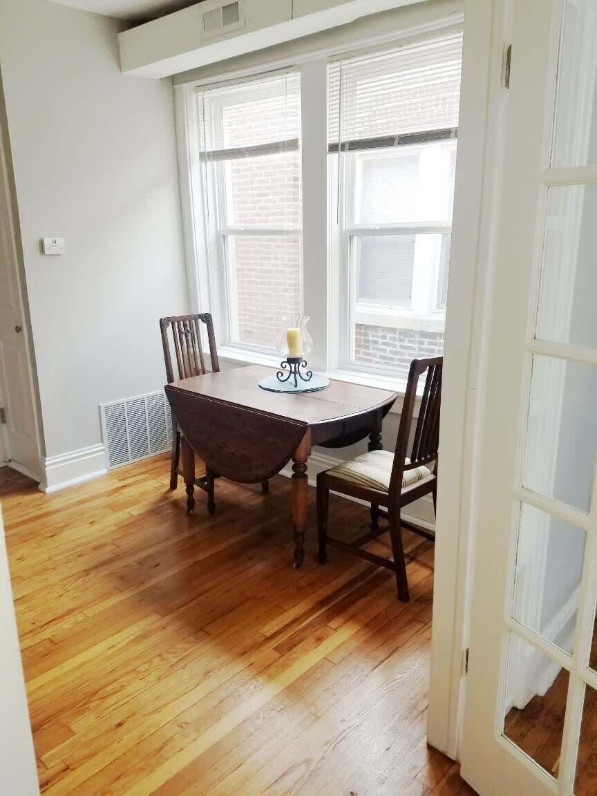 A dining area with a wooden table and chairs, large windows, and a radiator.