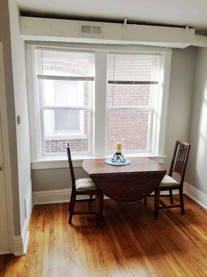 Un coin repas avec une table et deux chaises, une fenêtre avec des stores et un mur en briques.