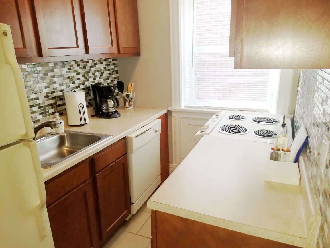 Une cuisine avec des meubles de rangement en bois, un pare-feu en carrelage, une cuisinière et une fenêtre avec des stores.