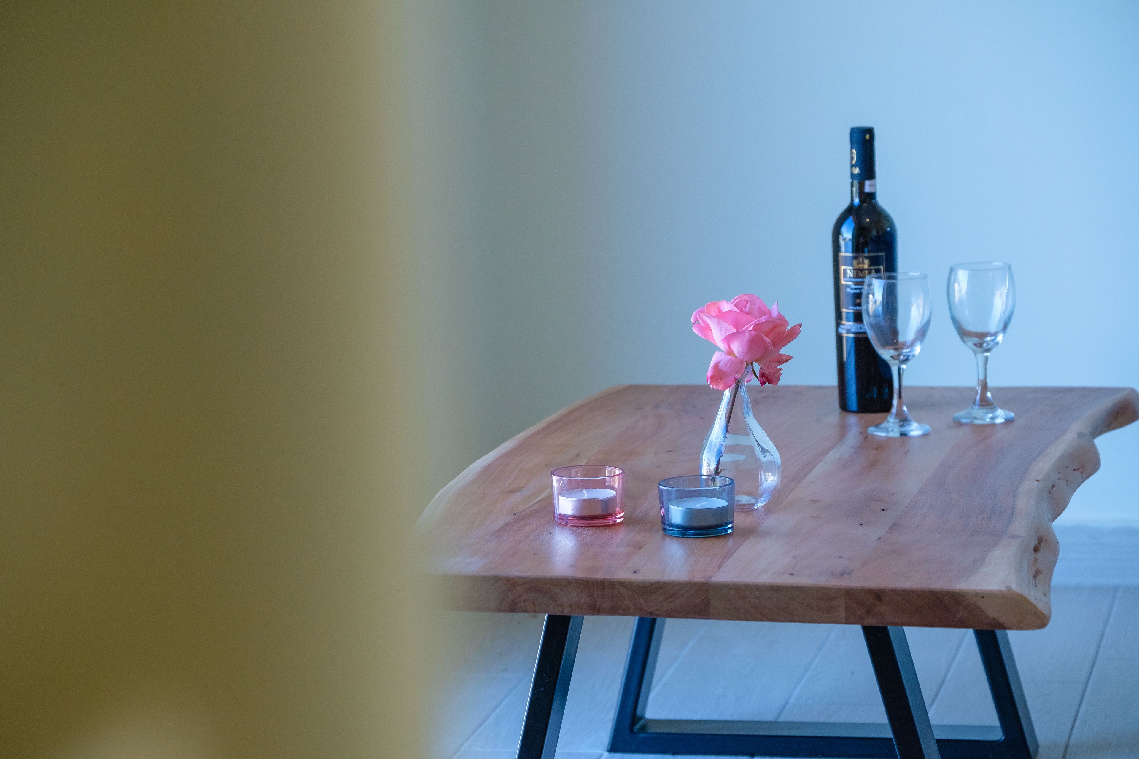 Een houten tafel met een fles wijn, twee glazen, een roze kaars en een kleine vaas met een roze bloem.
