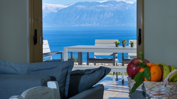 Una habitación con vista al mar y a las montañas, una mesa blanca con sillas y una cesta de fruta sobre una mesa.