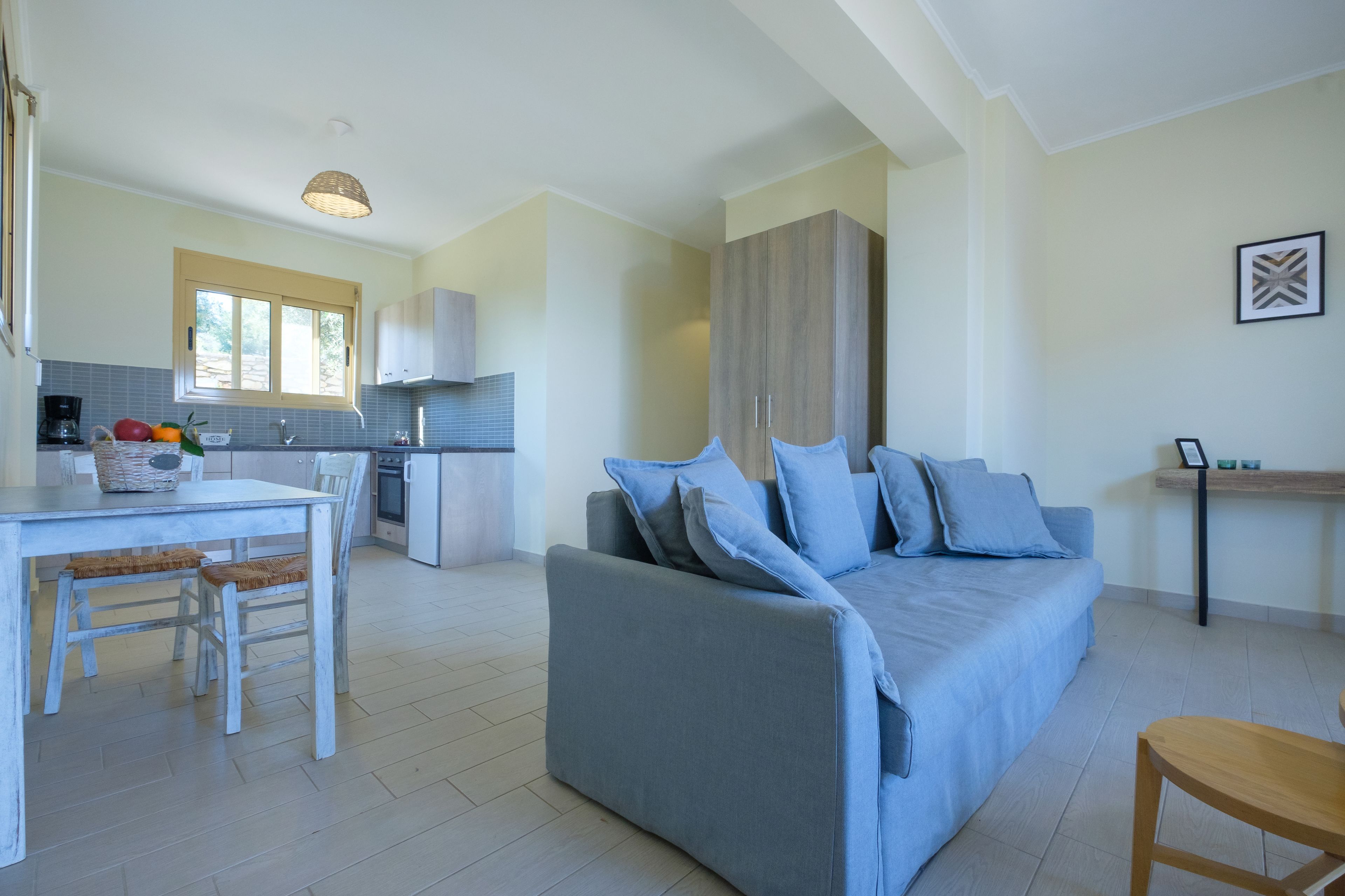 A modern living room with a blue sofa, a dining table with chairs, and a kitchen area with a window.