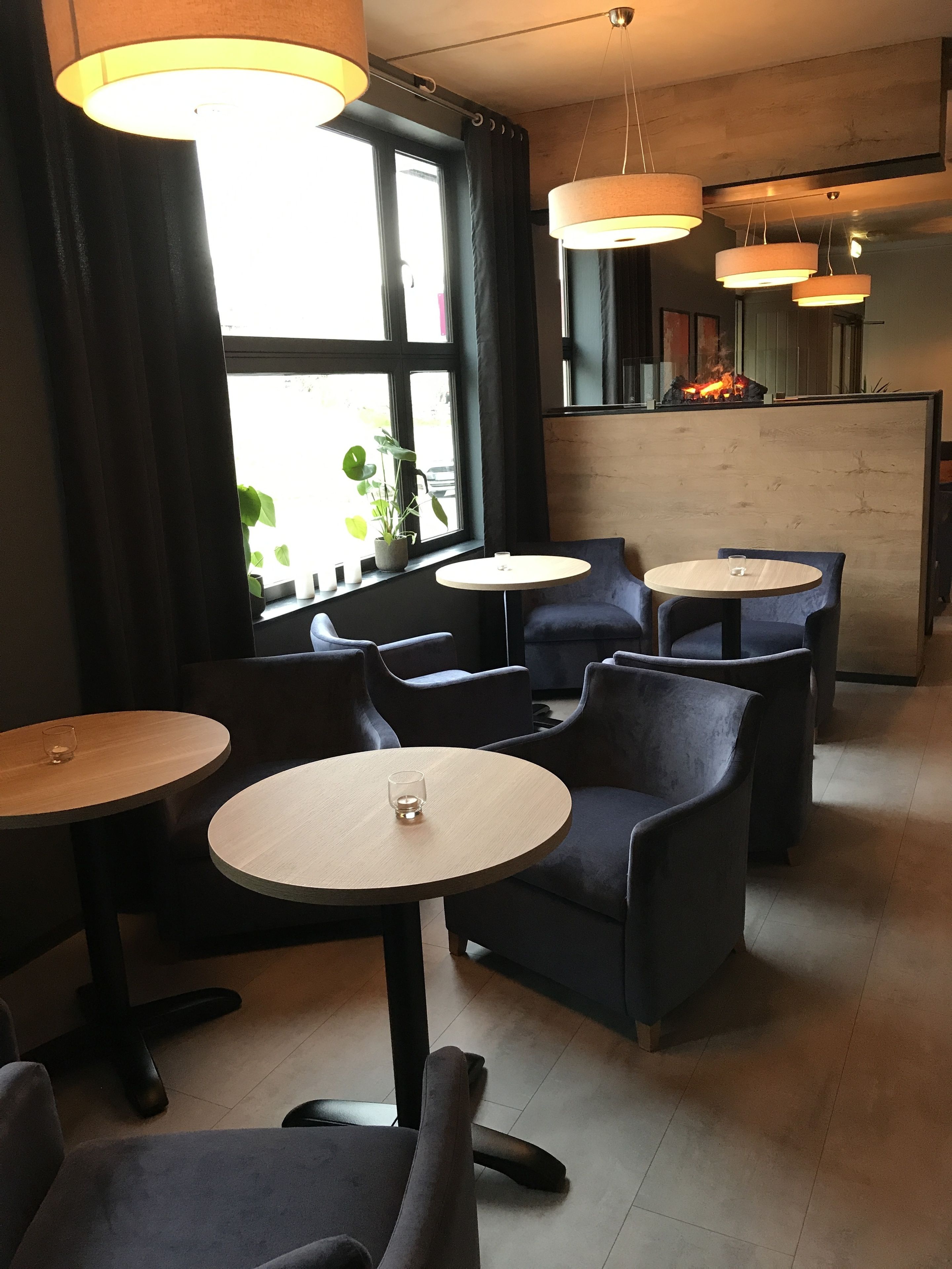 Ein modernes Interieur mit runden Tischen und dunklen Stühlen, großen Fenstern mit Vorhängen und einem sichtbaren Kamin.