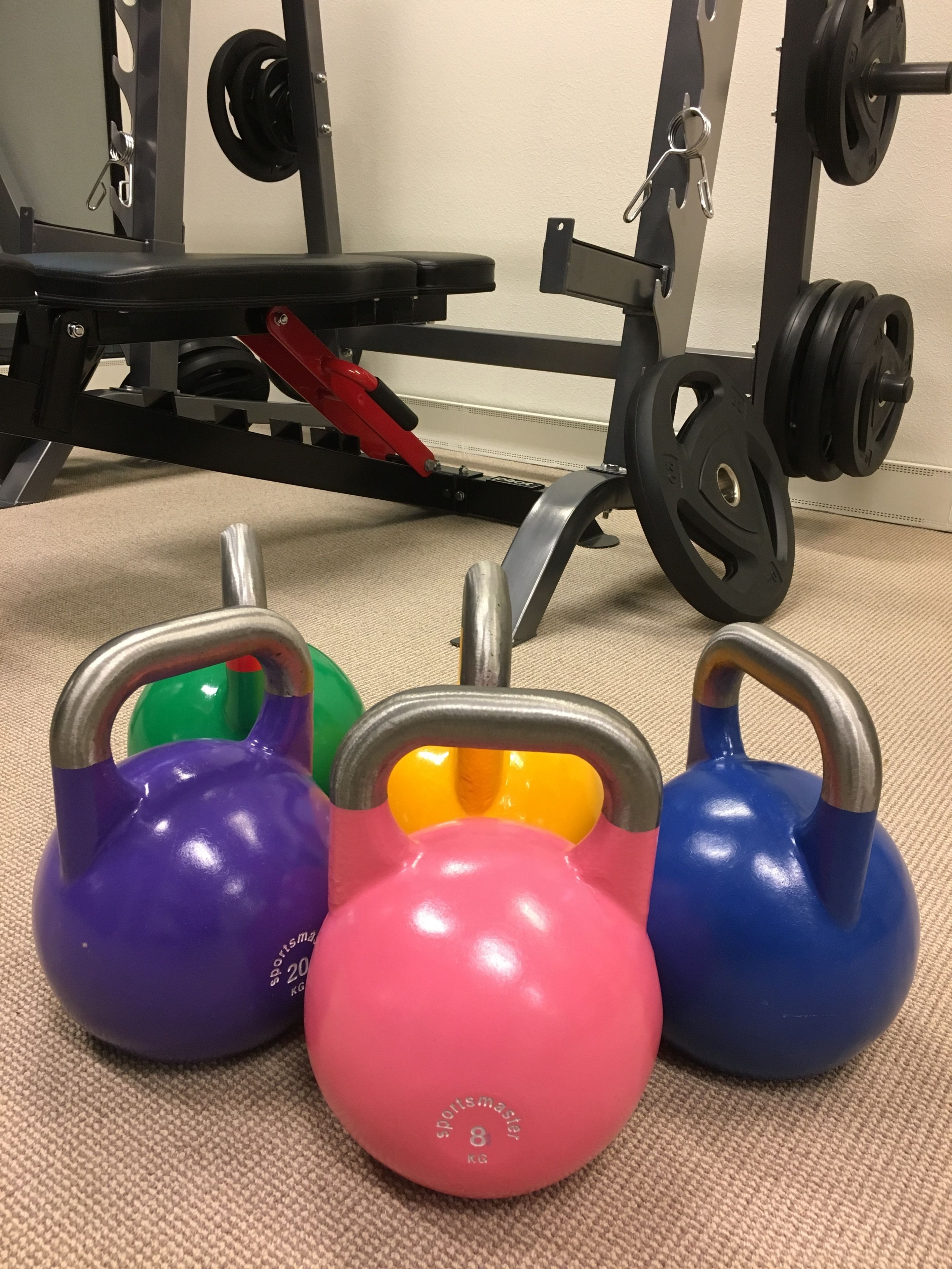 A set of colorful kettlebells on a carpeted floor with a weightlifting bench and barbell in the background.