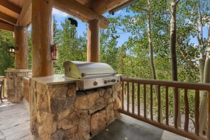 An outdoor grill area with stone walls, a wooden deck, and a view of trees.