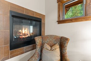 A fireplace with a modern design, a patterned armchair with a cushion, and a window with a view of greenery.