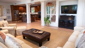 A spacious living room with a large TV, a leather ottoman, and a kitchen area in the background. - BREATHTAKING : on water front with panoramic views + pool + dock (Harbour Island)