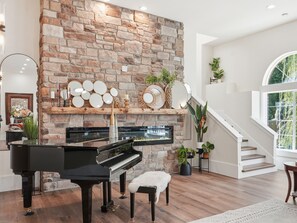 Un piano à queue se trouve dans une pièce spacieuse, dotée d’un mur en pierre, d’une cheminée et d’un escalier menant à un niveau supérieur.