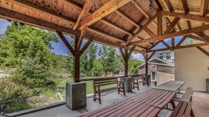 Un espacio al aire libre techado con bancos de madera y mesas de picnic, con vistas a un jardín ajardinado y una casa.