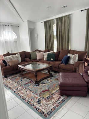 A living room with a brown sectional sofa, a glass-top coffee table, a patterned rug, and a large window with curtains. - Luxurious Private Estate with Hot Tub. Game Room, Grand Piano- Near Ski Resort (Salt Lake City)