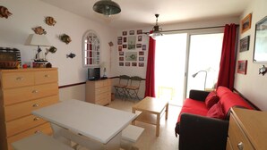 Un espacio habitable compacto con una mesa de comedor, un sofá, un escritorio y un aparador. Hay una ventana con cortinas rojas, un ventilador de techo y adornos en la pared, incluyendo cuadros y conchas.
