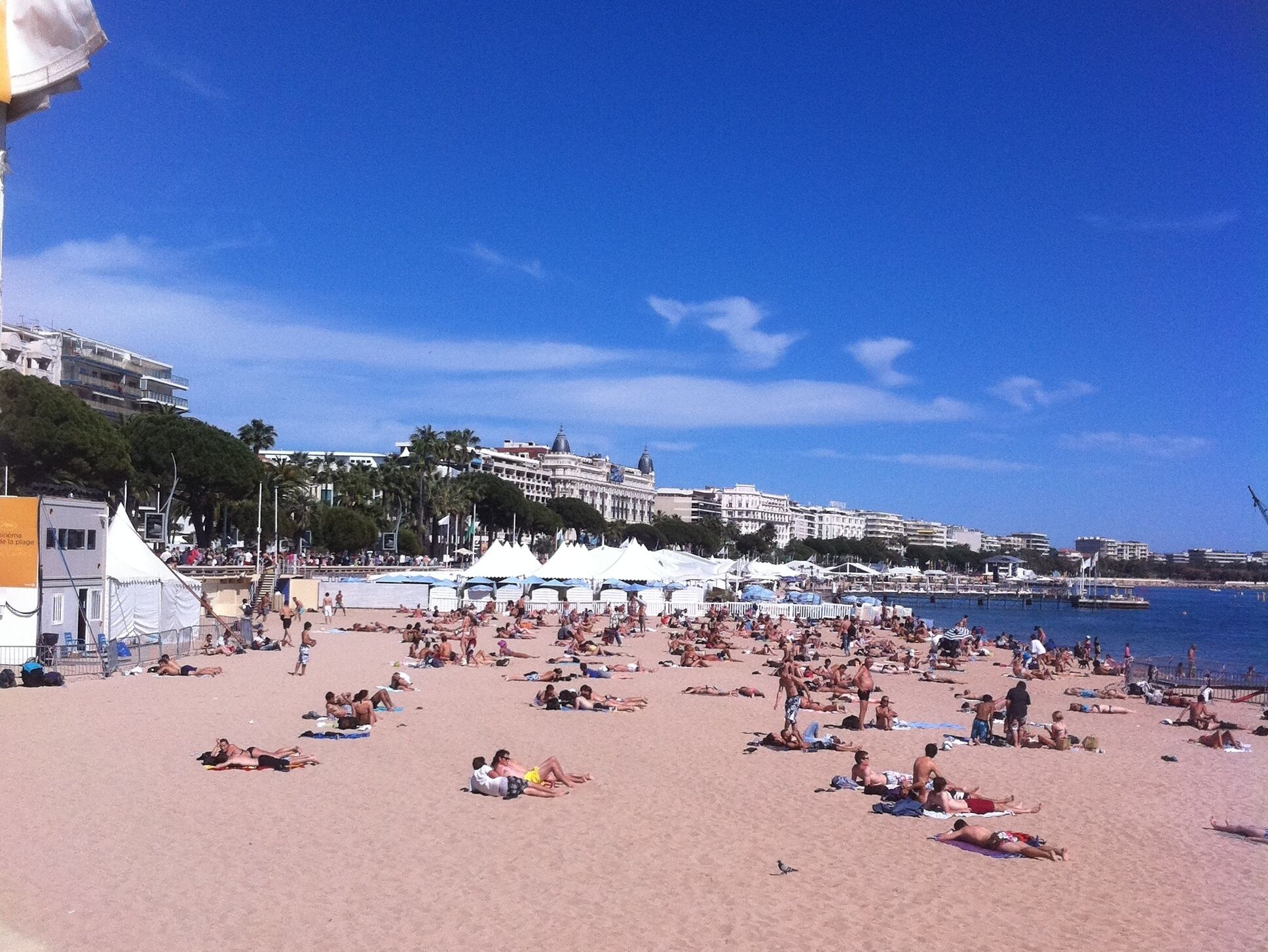 A crowded beach with people sunbathing and swimming, white tents, and a clear blue sky.
