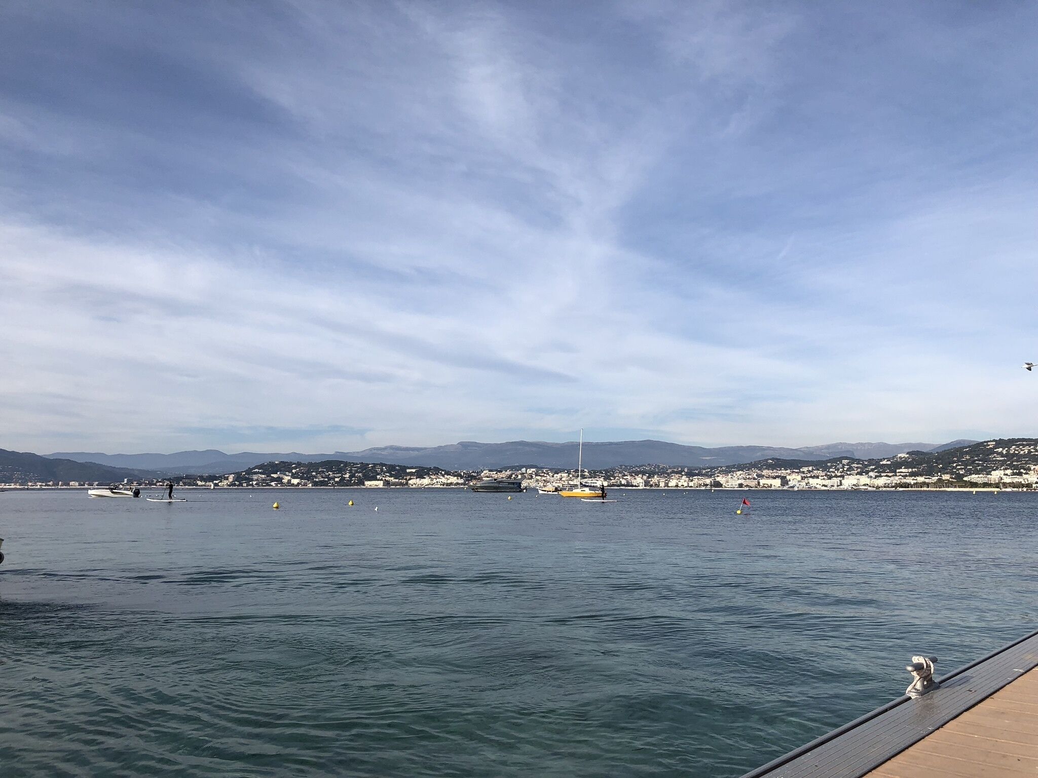 Vista costera con aguas tranquilas, un muelle y edificios a lo lejos.