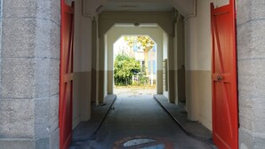 Ein Korridor mit roten Türen, einem Betonboden und Blick auf ein Gebäude mit blauen Fensterläden in der Ferne.