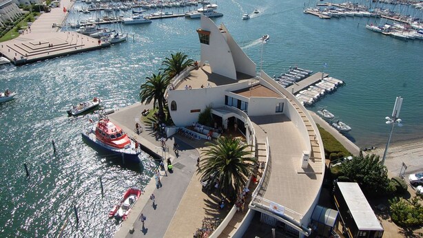 A marina with boats docked, a pedestrian walkway, and a building with a unique architectural design.