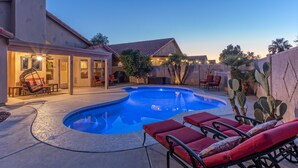 Un jardin avec une piscine, des chaises longues rouges, un patio couvert et des cactus.