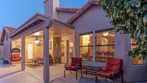Une terrasse ouverte avec deux chaises rouges et une table, un espace couvert avec un ventilateur de plafond, et une vue sur l’intérieur à travers de grandes fenêtres.