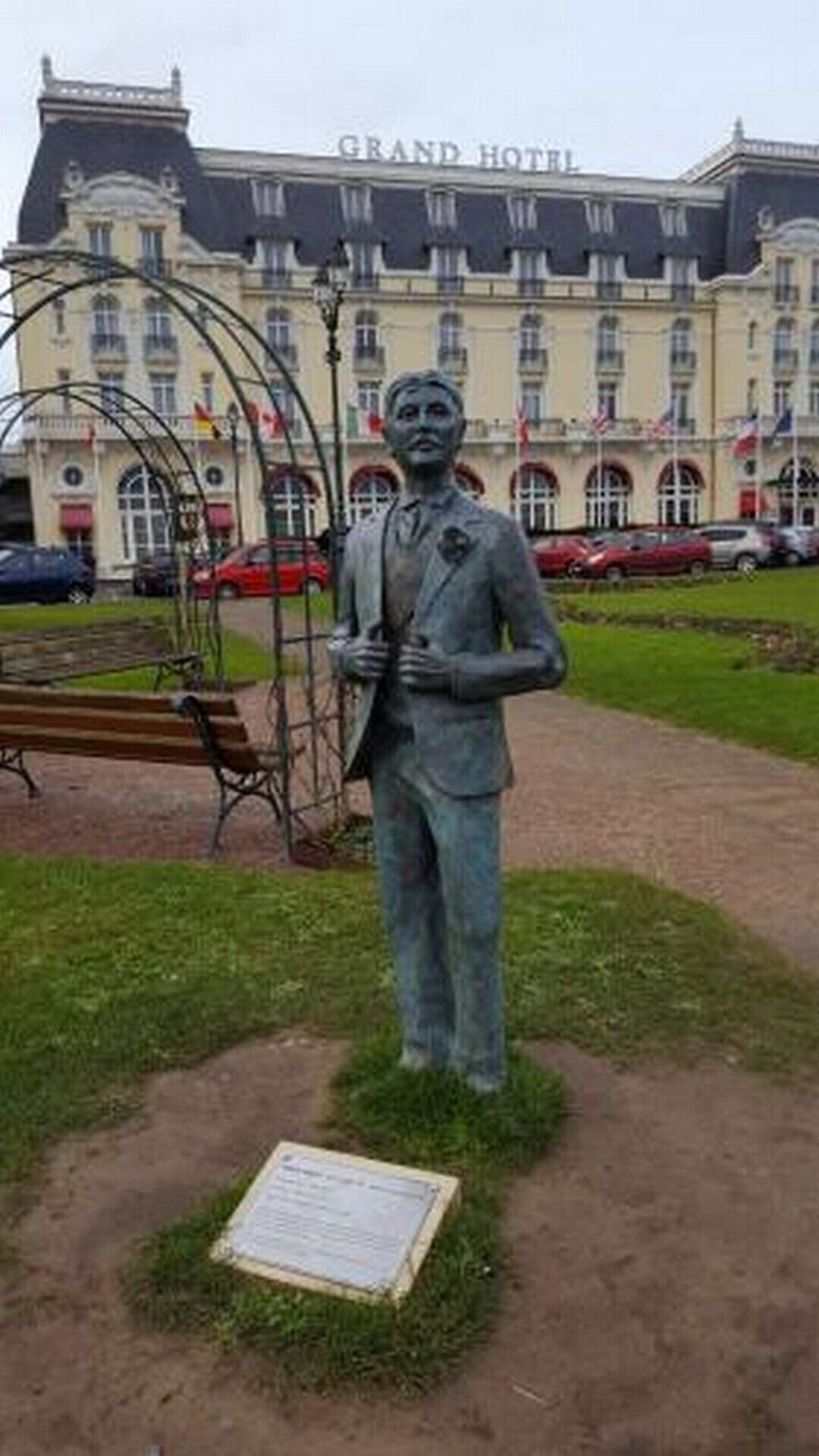 A statue in a park with a plaque in front of a building labeled "Grand Hotel".