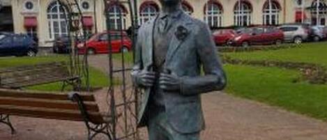 A statue in a park with a plaque in front of a building labeled "Grand Hotel".