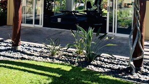Ein überdachter Außenbereich mit Glasveranda, umgeben von einem Garten mit Steinen und Pflanzen, Palmen und einem Gebäude mit Ziegeldach.