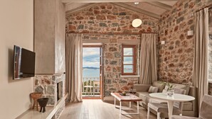 A spacious living room with a stone wall, a sofa, a round table, and a TV mounted on the wall. - 100 Rizes Seaside Resort - Small Luxury Hotels of The World (East Mani)
