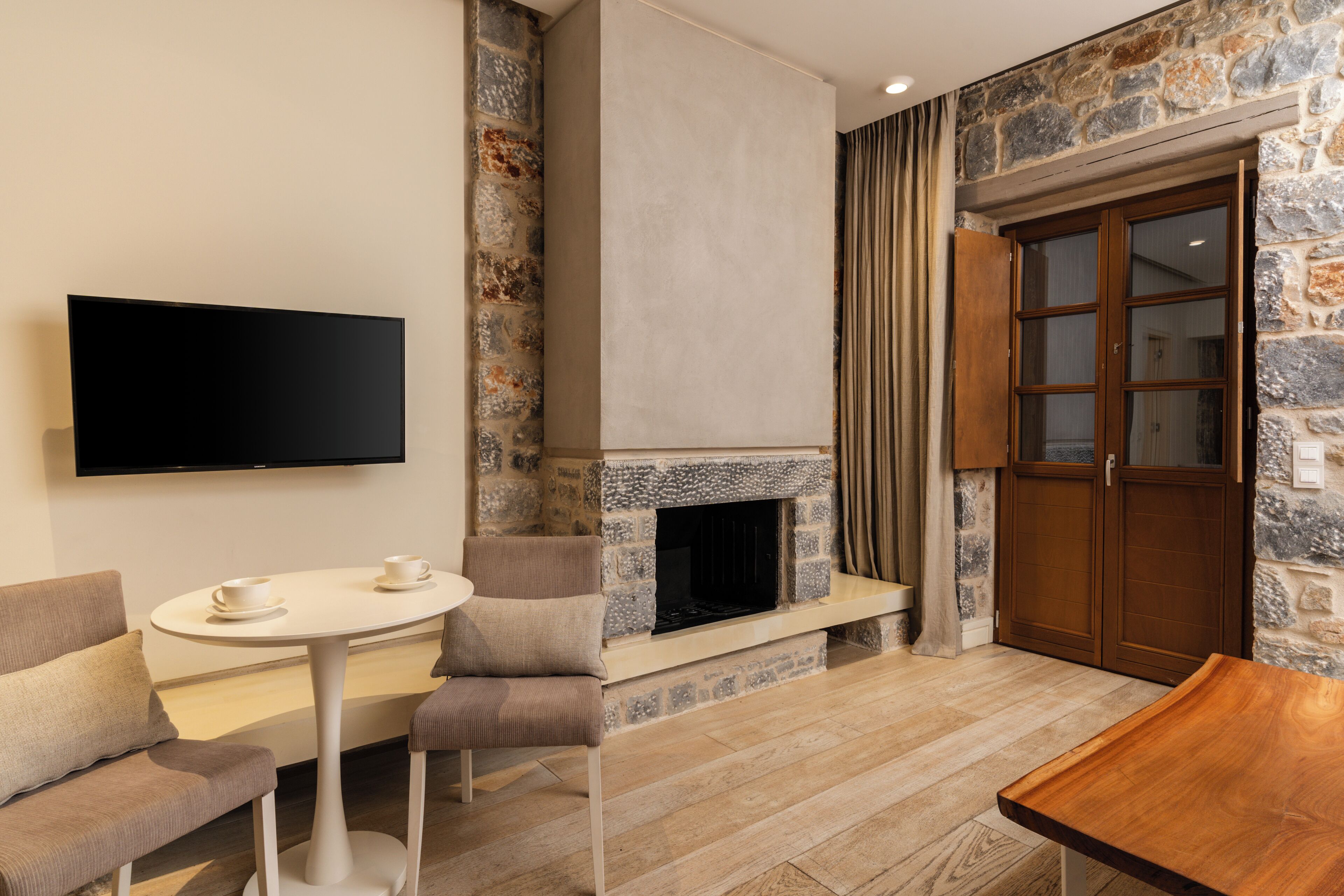 A modern living room with a stone fireplace, a flat-screen TV mounted on the wall, a round table with two chairs, and a wooden bench.