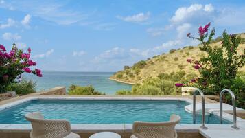 Ein Pool mit zwei Korbstühlen und einem kleinen Tisch mit Blick auf einen Strand und das Meer.