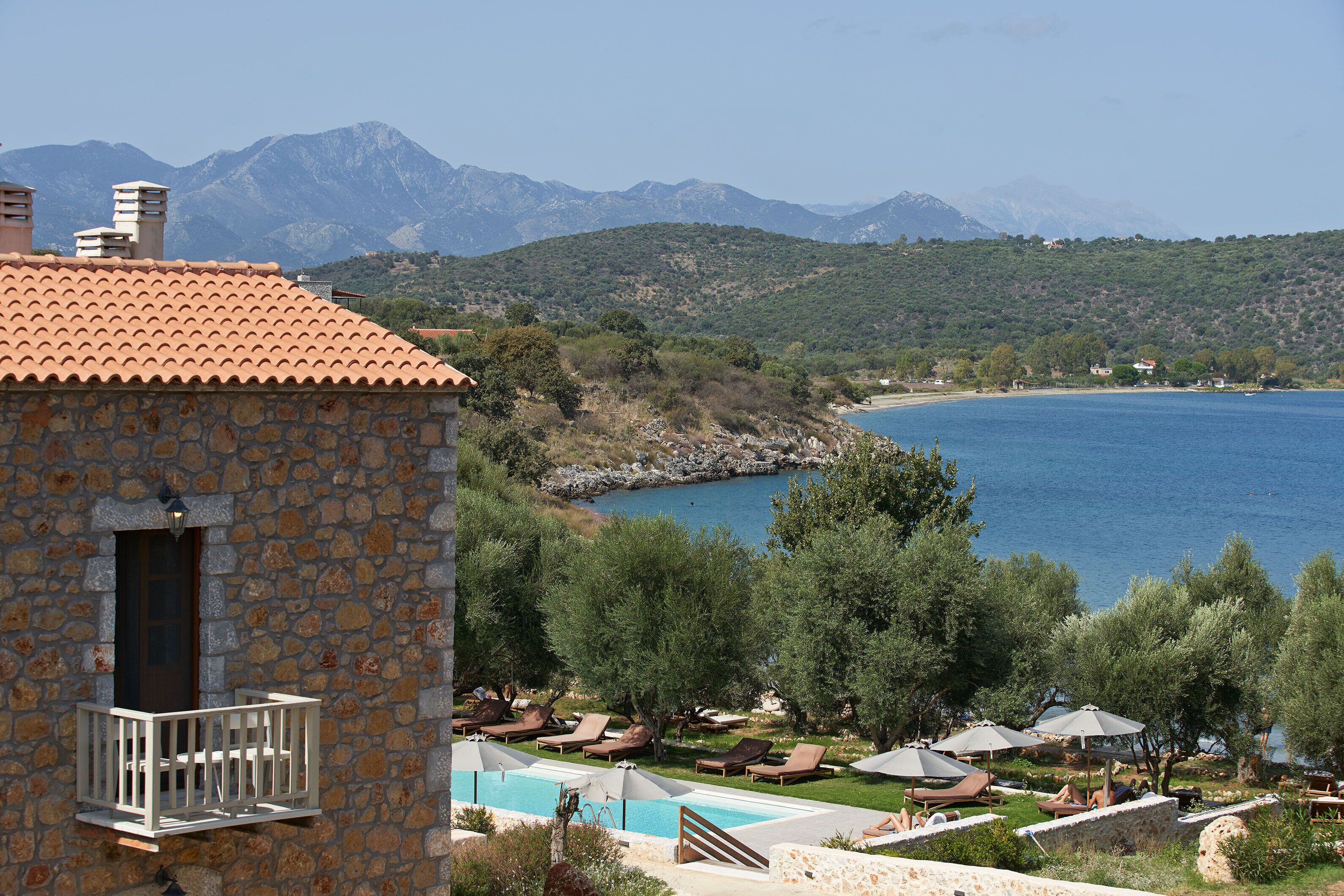 A stone building with a balcony, a swimming pool, and lounge chairs with umbrellas, overlooking a body of water and mountains.