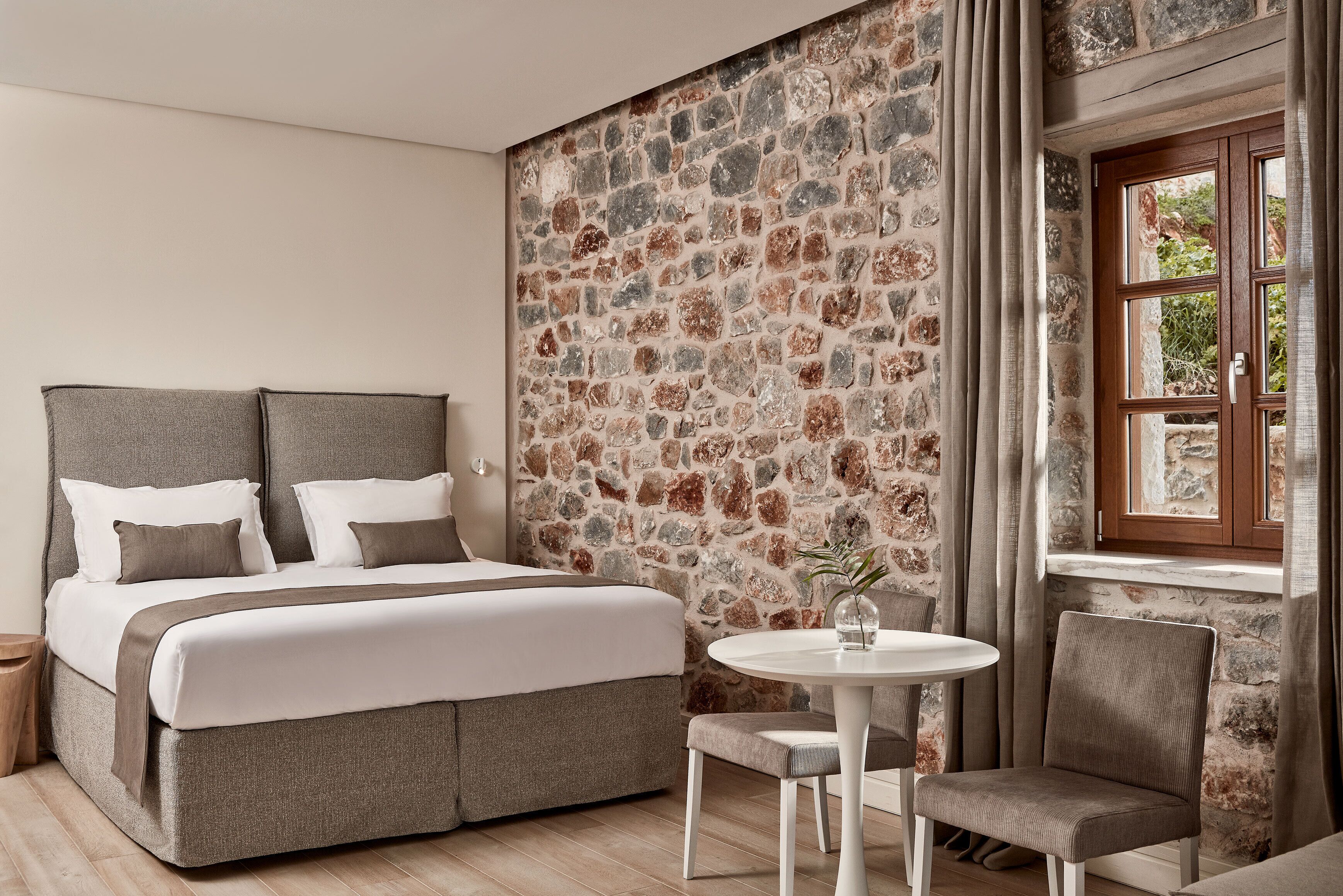 A bedroom with a stone wall, a bed, a round table, and a chair.