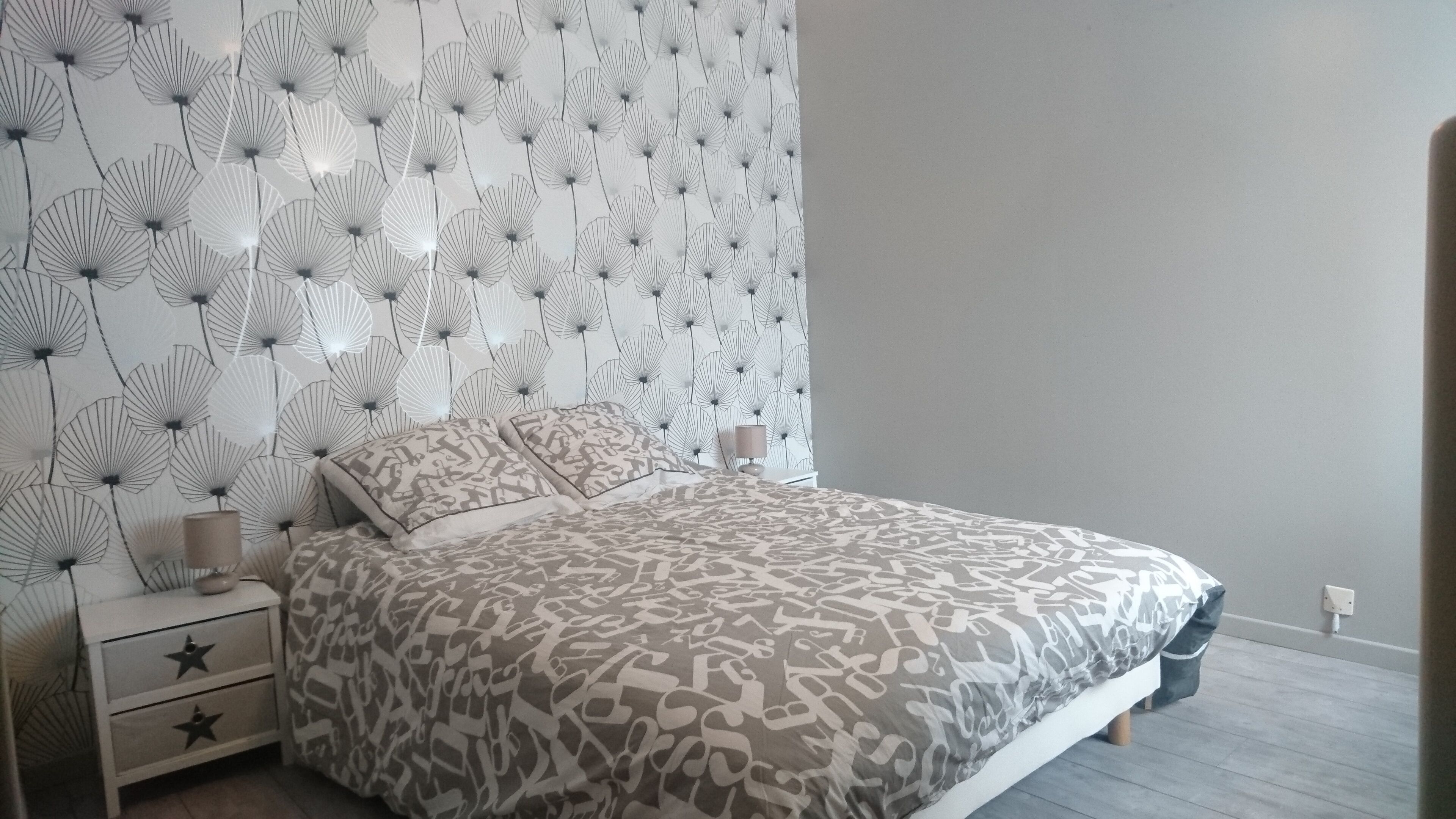 Ein Schlafzimmer mit einem bettenbezug mit Muster, einem Nachttisch mit Lampe und einer Wand mit Blättermotiv.