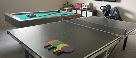 A room with a ping pong table, a pool table, and a television on the wall.