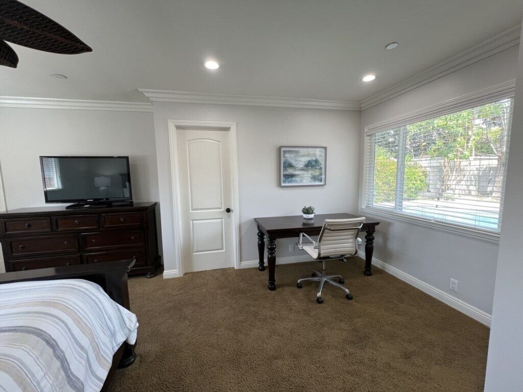 Un dormitorio con cama, escritorio, silla, televisor, puerta y ventana.