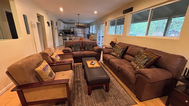 A spacious living room with a large brown sectional sofa, a wooden coffee table, and a fireplace. A kitchen is visible in the background, and there are large windows providing natural light. - Tahoe house with amazing mountain views (South Lake Tahoe)