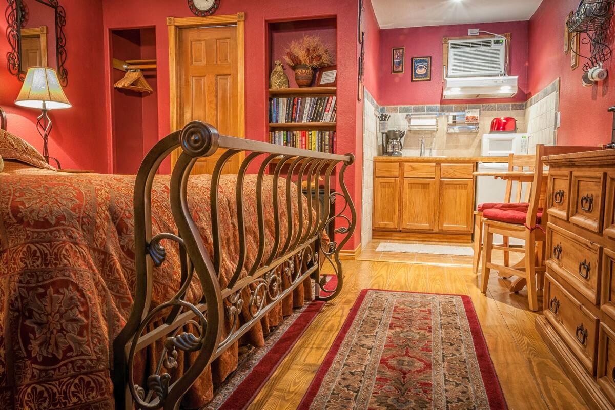 Un dormitorio con estructura de cama metálica, cómoda de madera y una zona de cocina con alacenas de madera y microondas.