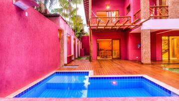 A pool area with a wooden deck and a red building with a balcony.