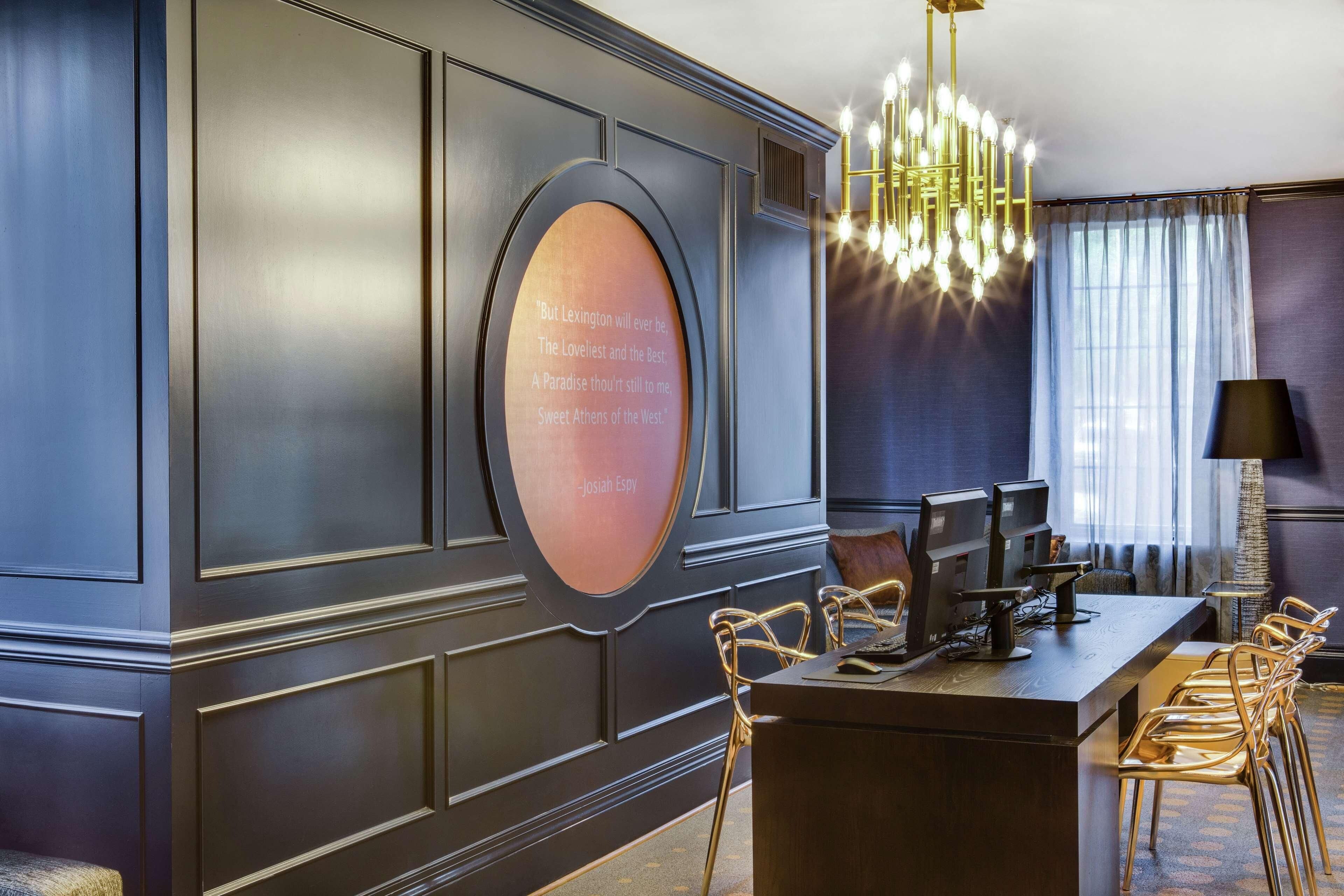 A modern hotel room with a dark wood panel wall, a round mirror with a quote, a chandelier, a desk with computers, and a seating area with chairs.