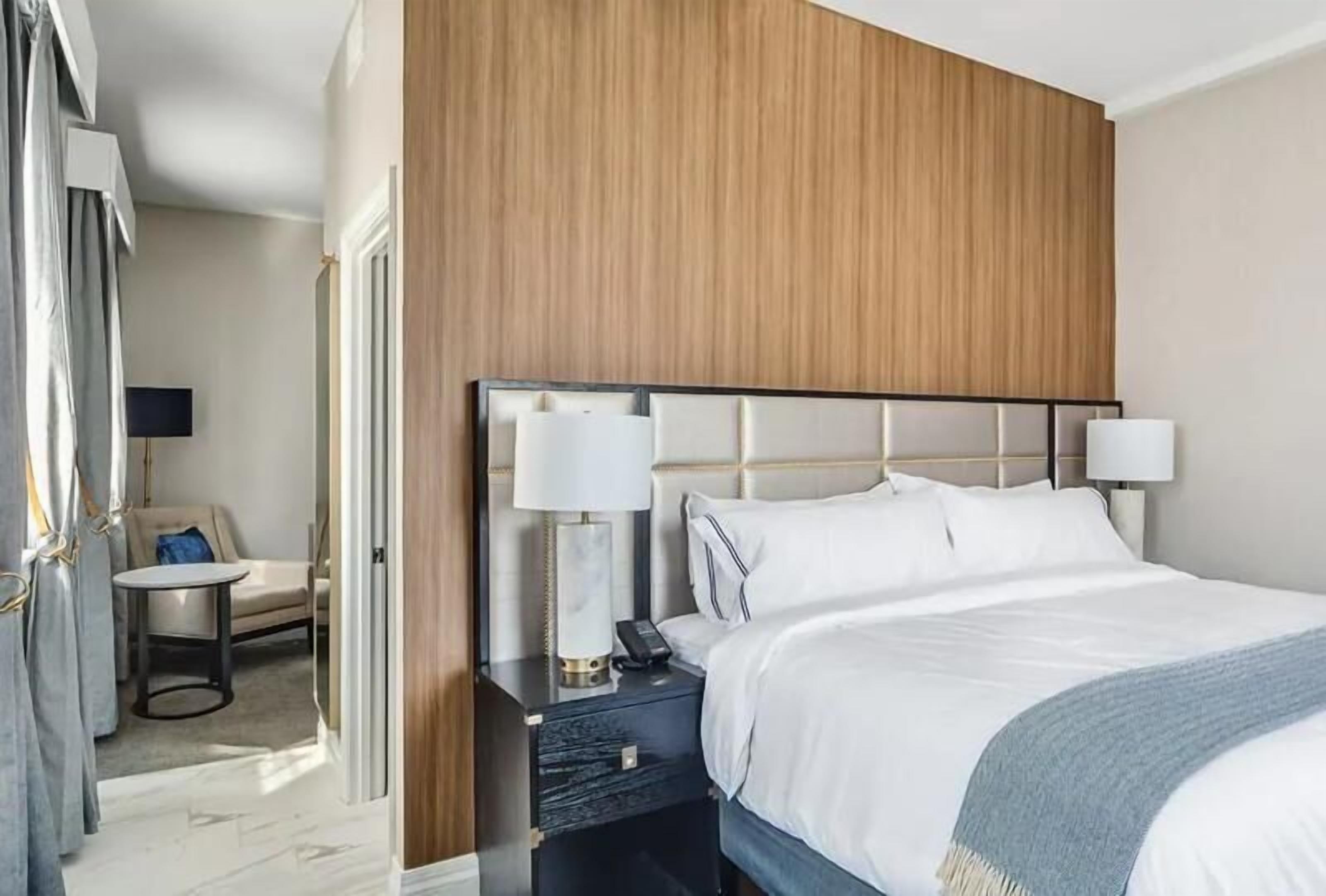 A hotel room with a large bed, two bedside lamps, a small table with a chair, and a TV.