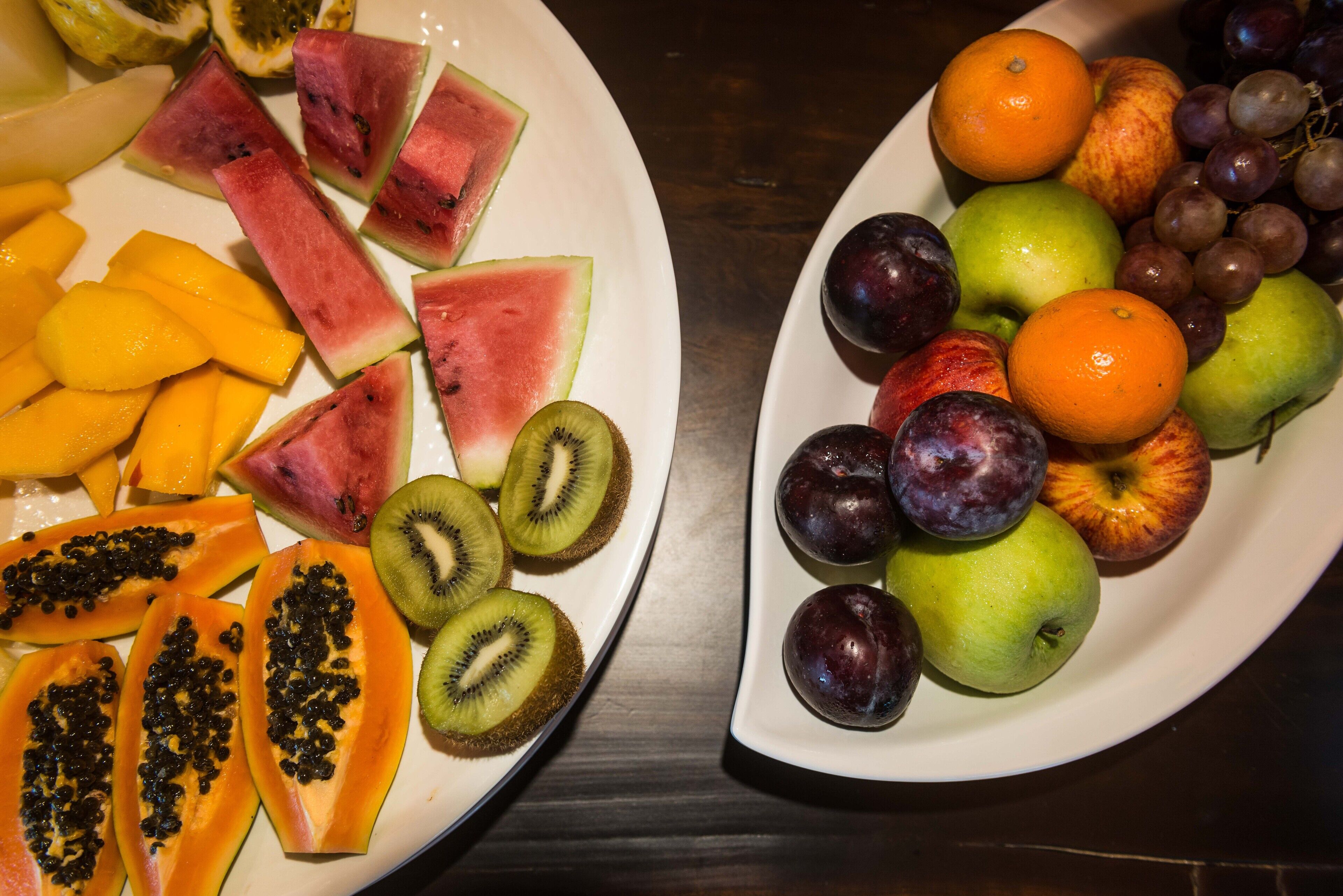 Two plates of assorted fruits including watermelon, kiwi, papaya, mango, apples, plums, oranges, and grapes.